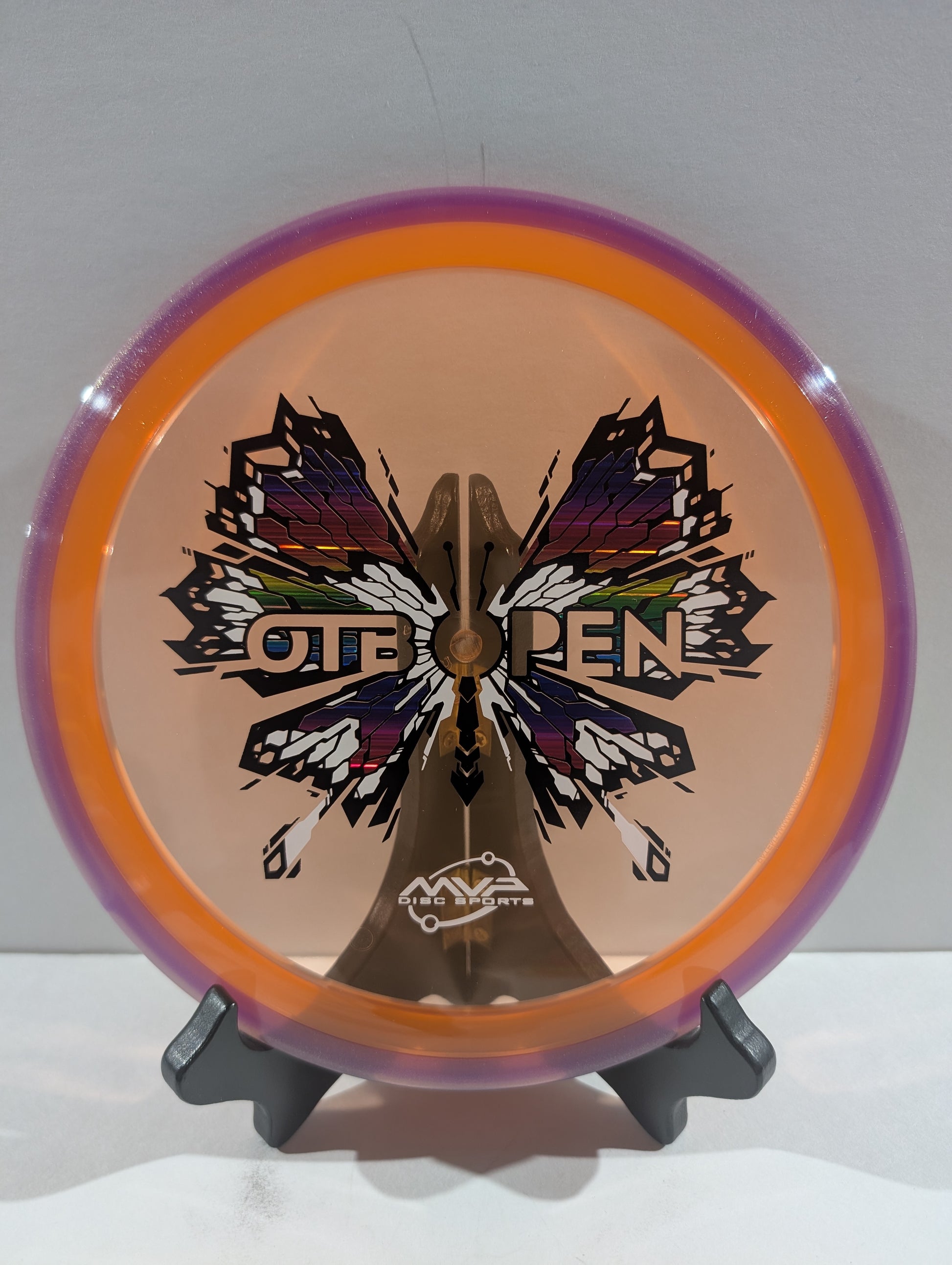 Disc golf disc with butterfly design and 'OTB Open' branding on a stand against a white background
