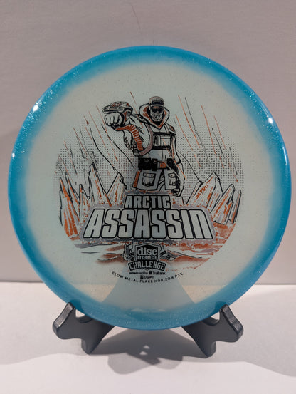 A Discmania Arctic Assassin disc golf disc with a blue rim and a white center featuring a graphic of an Arctic soldier and mountains, displayed on a small stand.