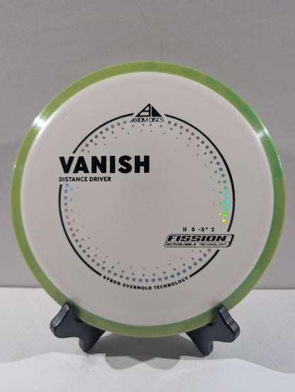 A disc golf disc on a small display stand, featuring a white flight plate with a light green rim, black printed graphics, and the text 'VANISH' and 'Distance Driver'.