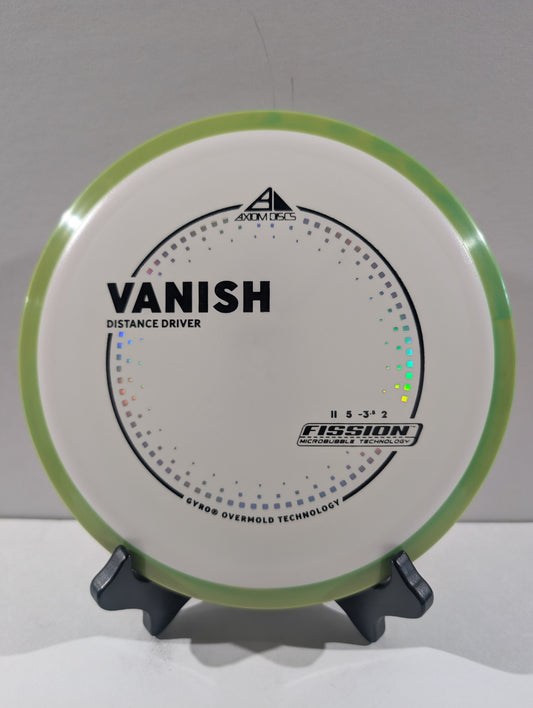 A disc golf disc on a small display stand, featuring a white flight plate with a light green rim, black printed graphics, and the text 'VANISH' and 'Distance Driver'.