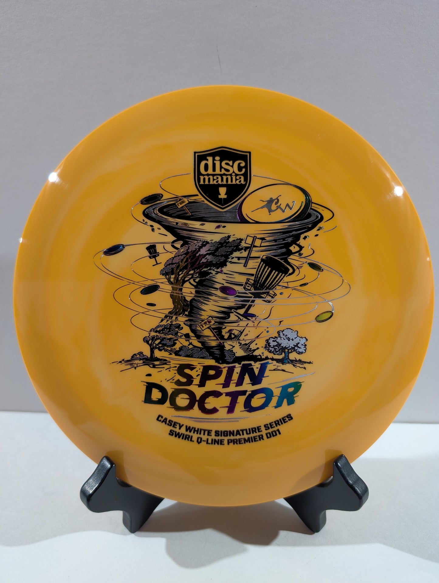 An orange Discmania Spin Doctor flying disc displayed on a small black stand against a neutral gray background.