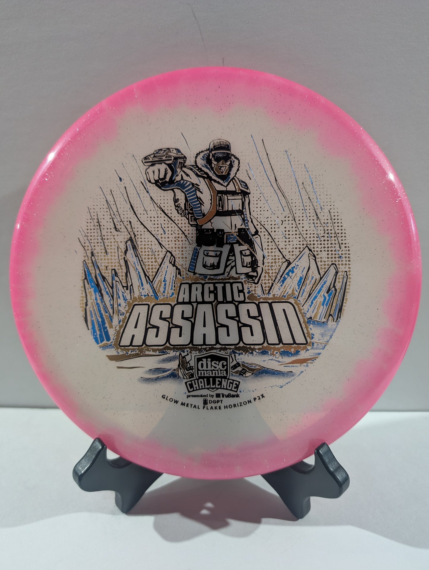 A Discmania Arctic Assassin disc golf disc with a pink rim and a white center featuring a graphic of an Arctic soldier and mountains, displayed on a small stand.