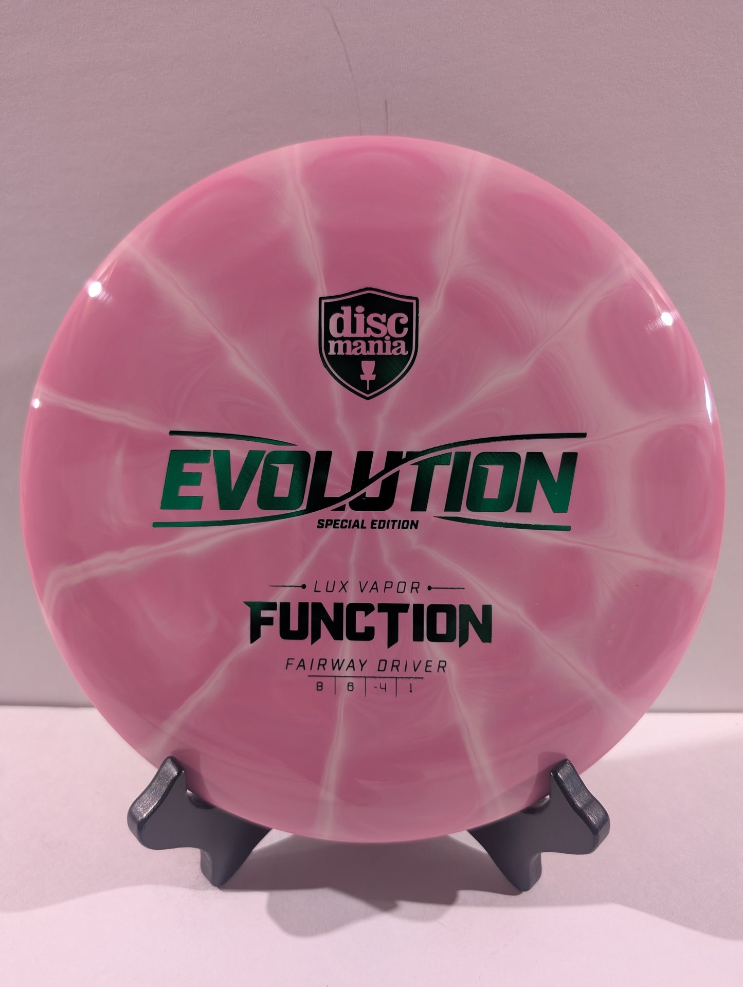A pink swirled Discmania Evolution Lux Vapor Function fairway driver disc golf disc on a black display stand against a neutral gray background.