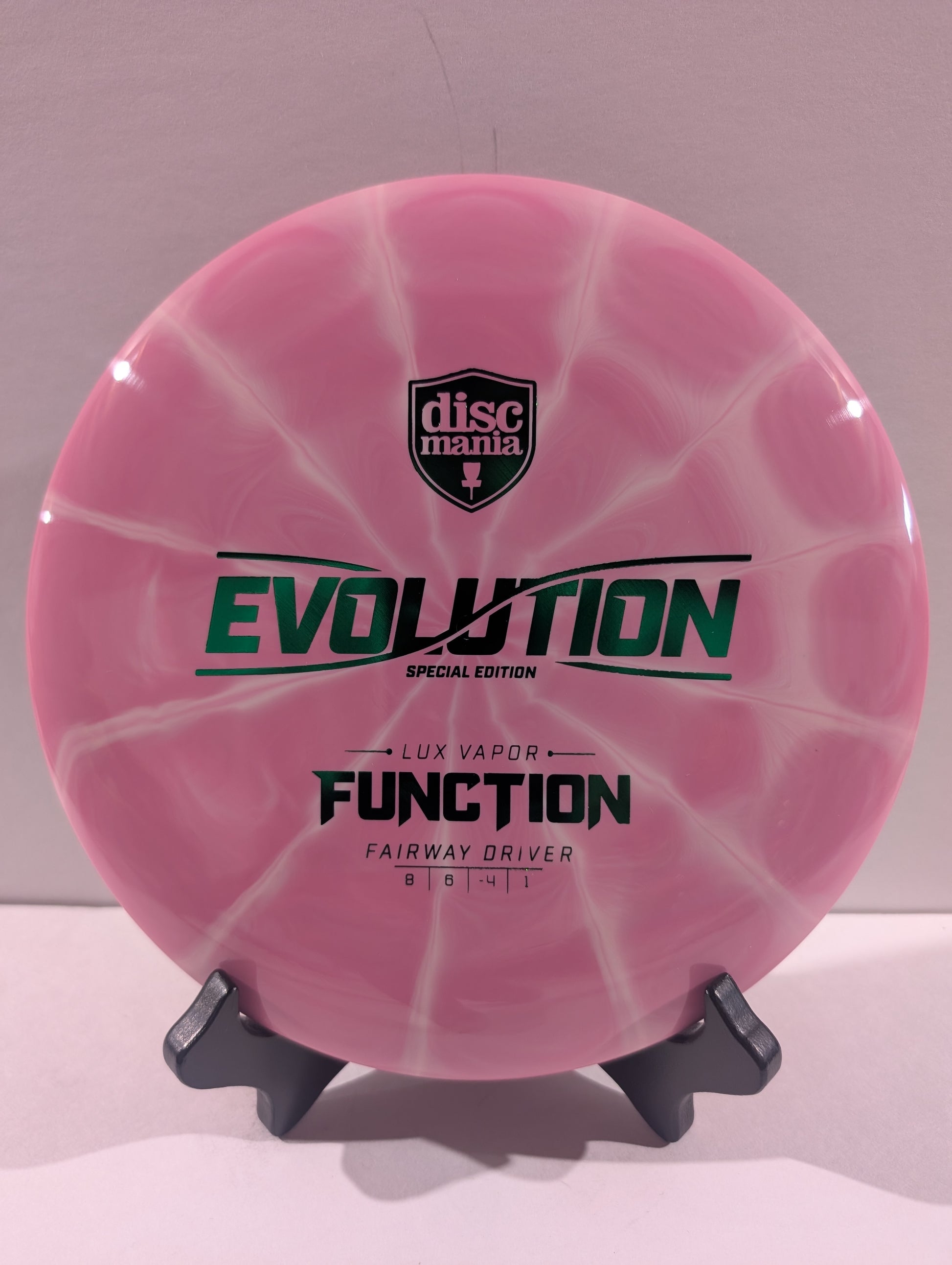 A pink swirled Discmania Evolution Lux Vapor Function fairway driver disc golf disc on a black display stand against a neutral gray background.