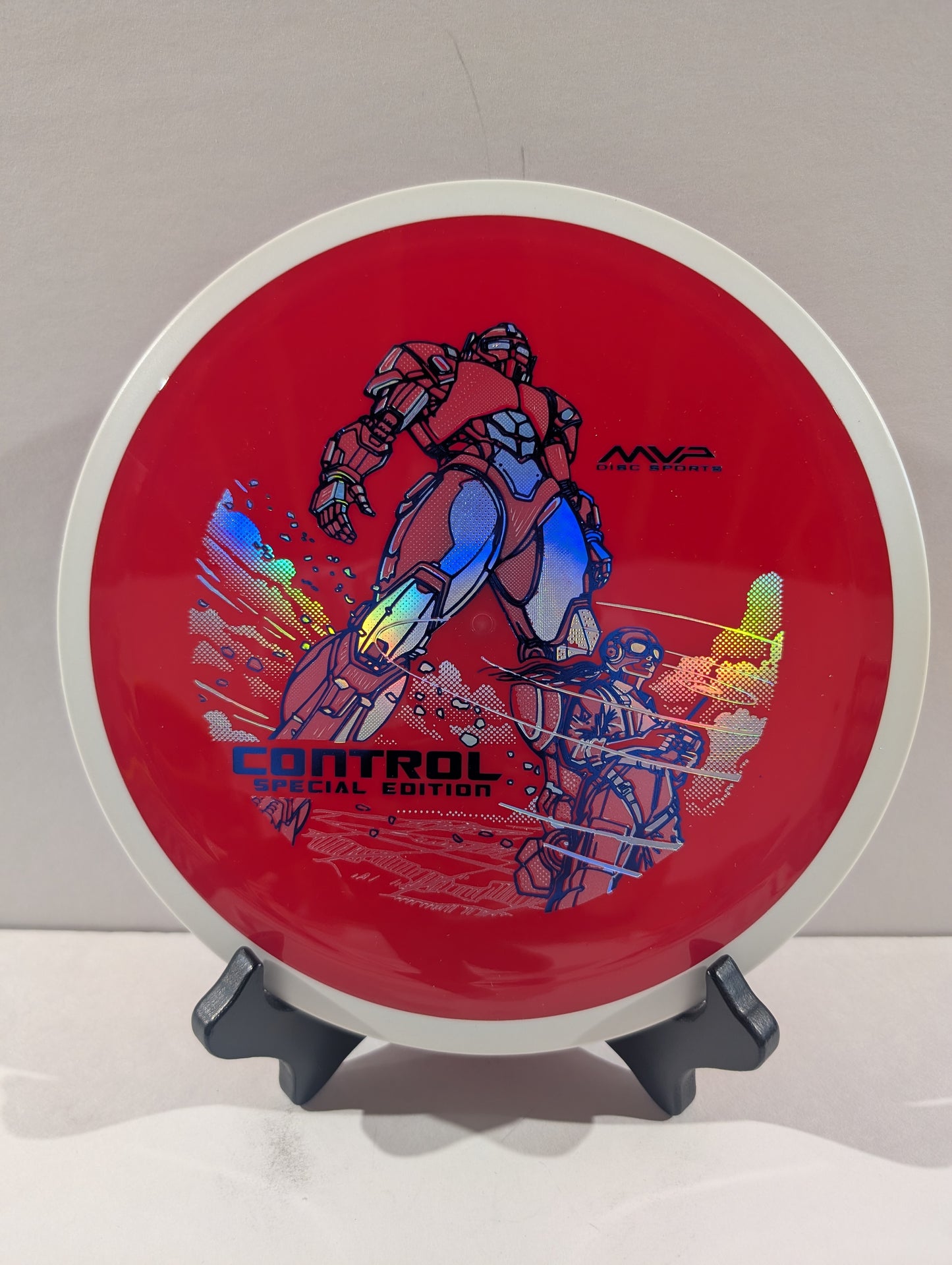 A red disc golf disc with a white outer rim, featuring a blue and black robot graphic and holographic foil accents, displayed on a small gray stand against a neutral light gray background.
