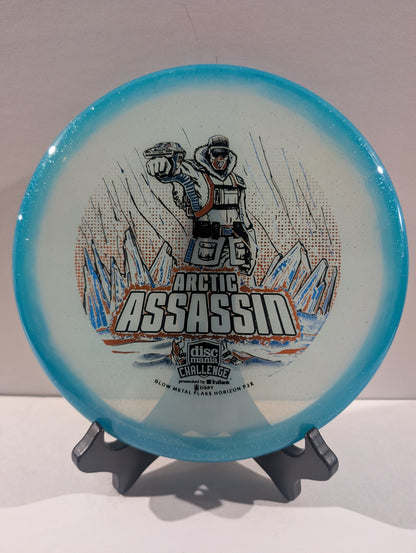 A Discmania Arctic Assassin disc golf disc with a blue rim and a white center featuring a graphic of an Arctic soldier and mountains, displayed on a small stand.
