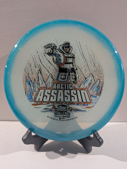 A Discmania Arctic Assassin disc golf disc with a blue rim and a white center featuring a graphic of an Arctic soldier and mountains, displayed on a small stand.
