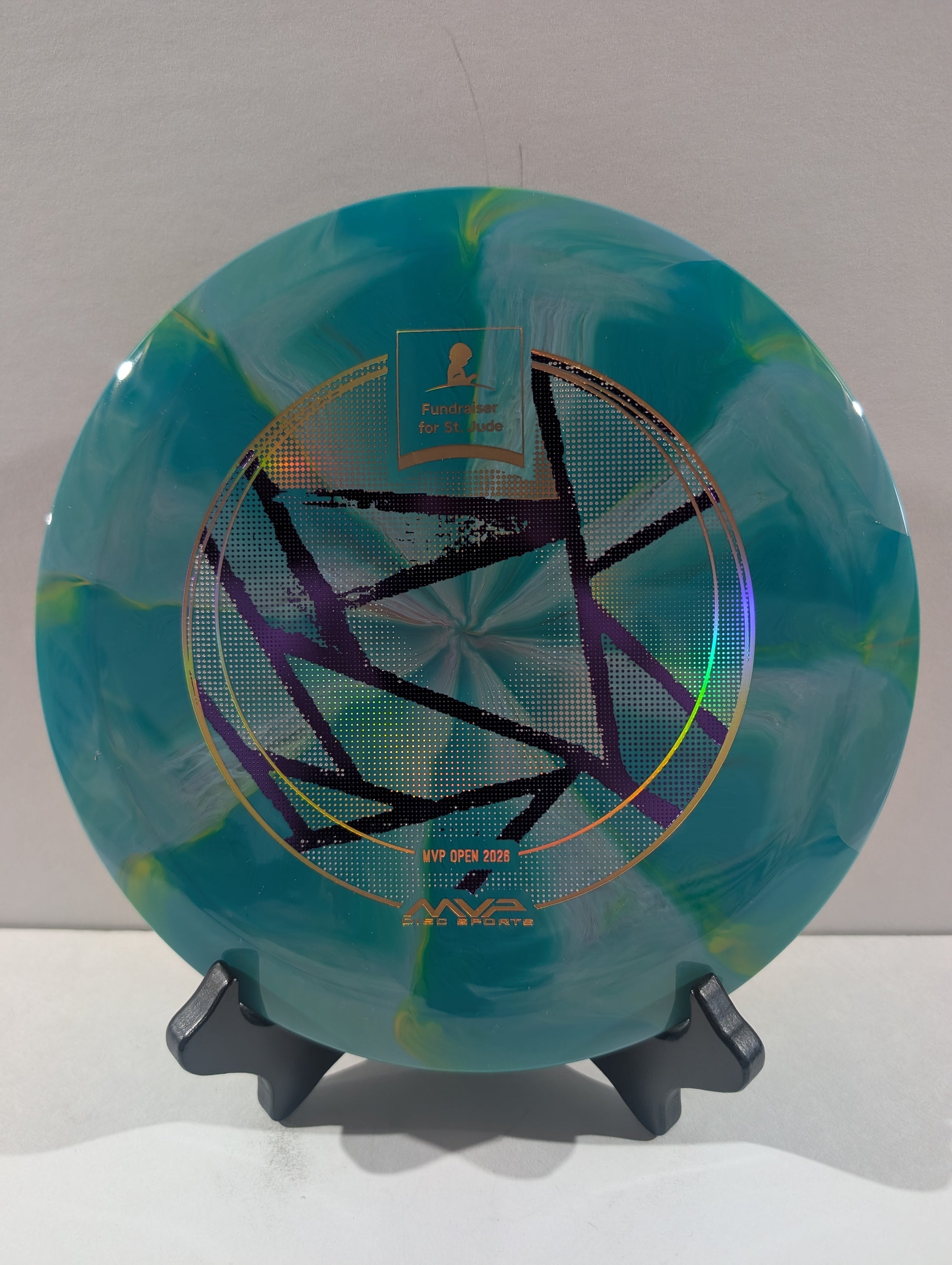 A disc golf disc with a teal and green marbled pattern, featuring a black geometric star design and foil accents, displayed on a black stand against a neutral background.