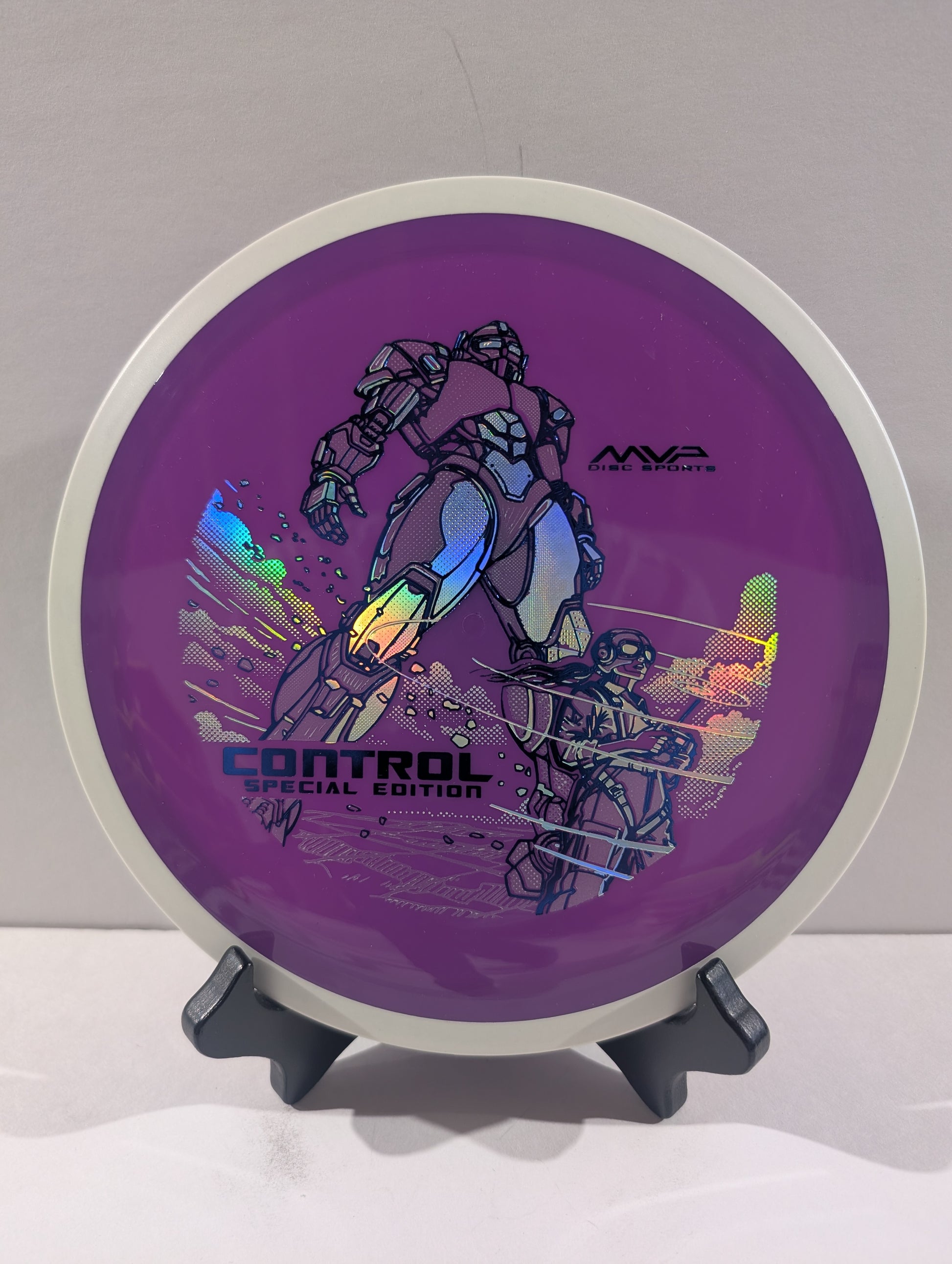 A purple disc golf disc with a white outer rim, featuring a graphic of a mecha robot and the text 'CONTROL SPECIAL EDITION' and 'MVP'. The disc is displayed on a small black stand against a neutral light gray background.