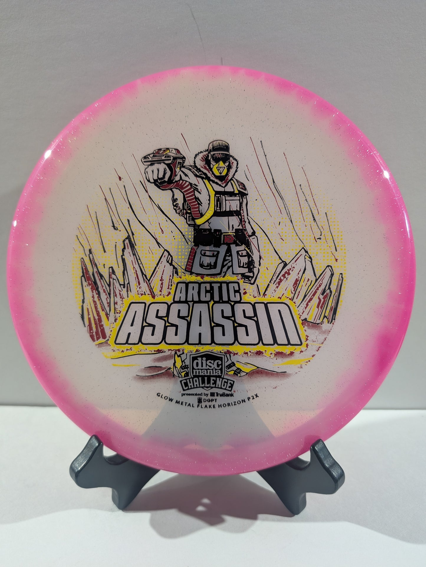 A Discmania Arctic Assassin disc golf disc with a pink rim and a white center featuring a graphic of an Arctic soldier and mountains, displayed on a small stand.