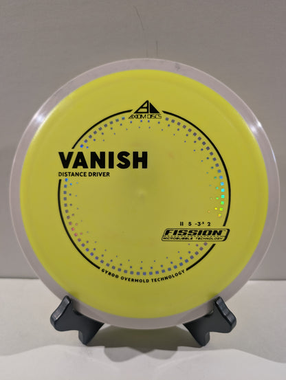 A yellow disc golf disc, specifically the Vanish distance driver in Fission, displayed on a small black stand against a neutral gray background.