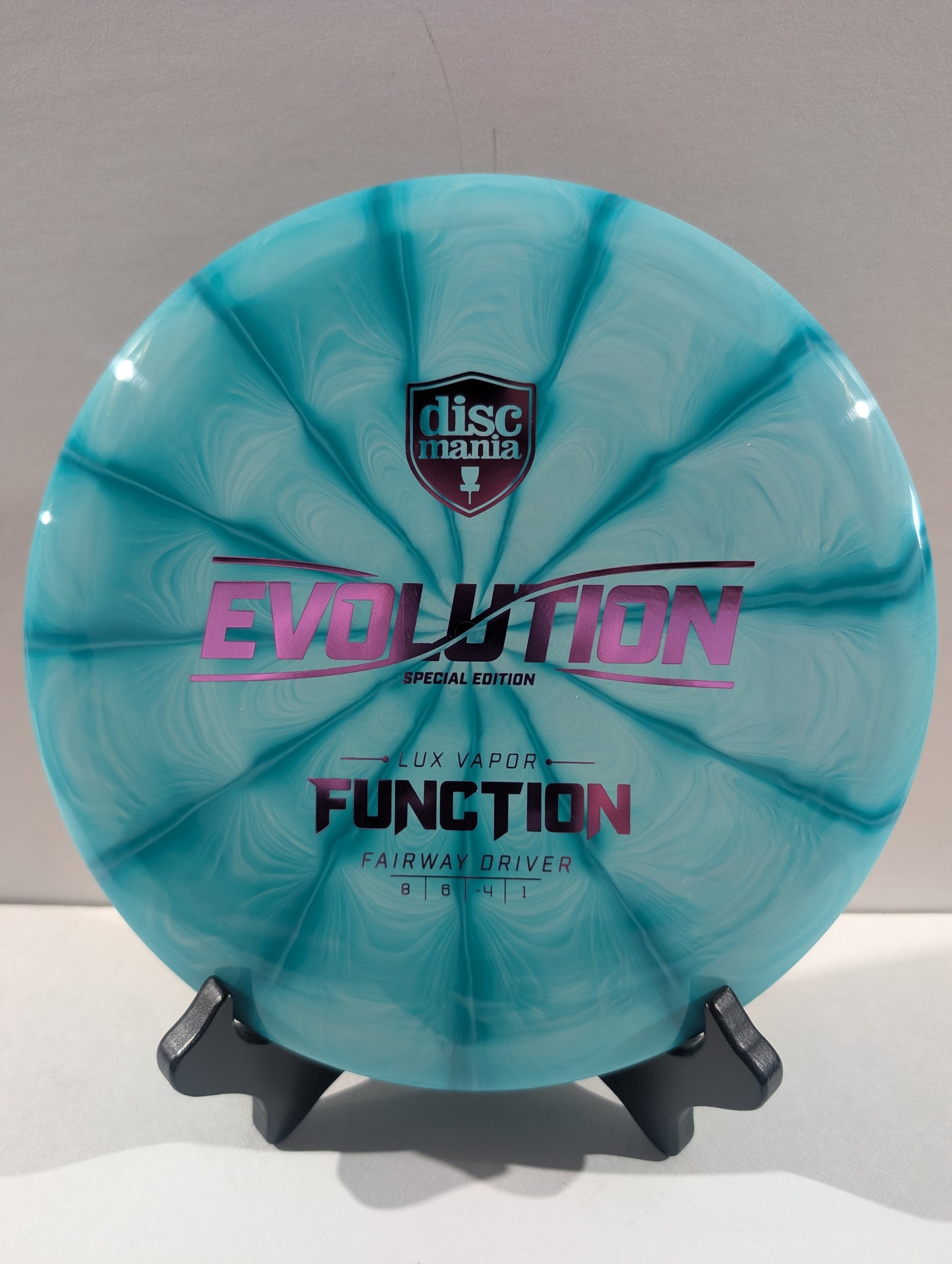 A turquoise swirled Discmania Evolution Lux Vapor Function fairway driver disc golf disc on a black display stand against a neutral gray background.