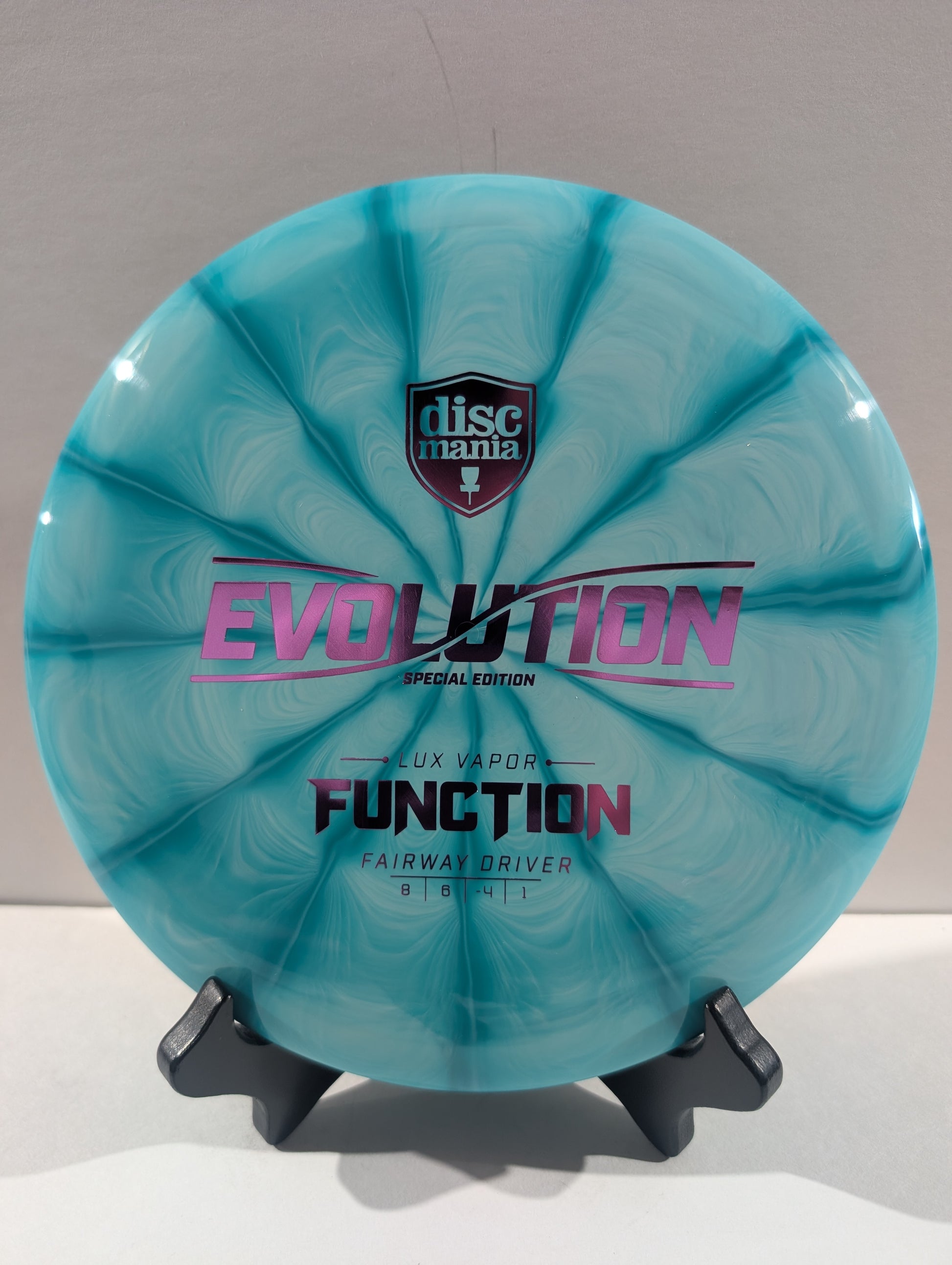 A turquoise swirled Discmania Evolution Lux Vapor Function fairway driver disc golf disc on a black display stand against a neutral gray background.