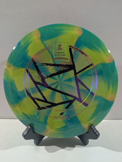 A disc golf disc with a green and yellow marbled pattern, featuring a geometric star design in the center and a foil stamp, displayed on a black stand against a neutral background.