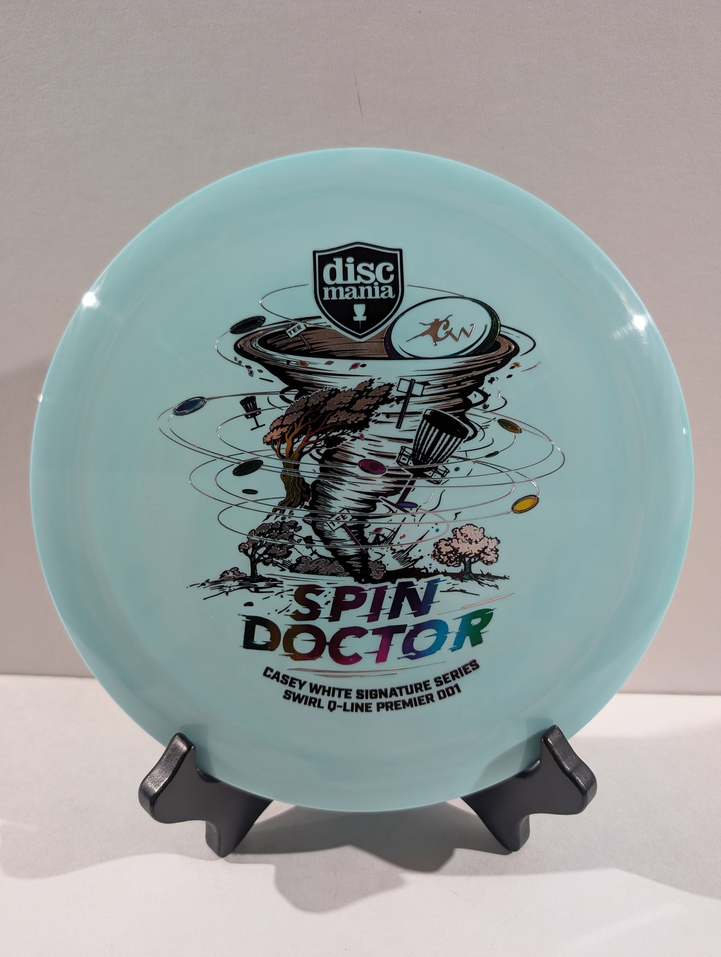 A light blue Discmania Spin Doctor disc golf disc displayed on a black stand against a neutral gray background.