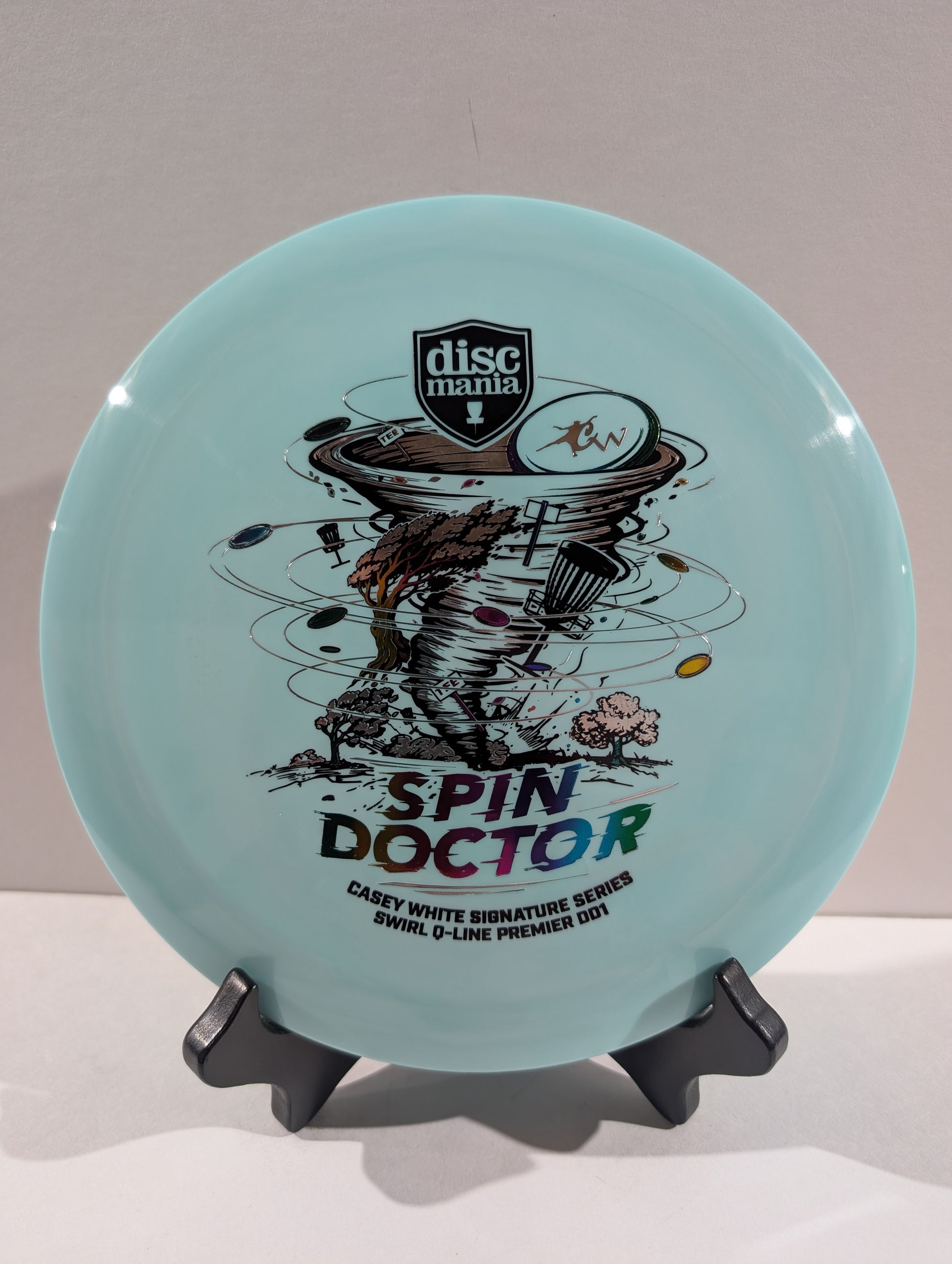 A light blue Discmania Spin Doctor disc golf disc displayed on a black stand against a neutral gray background.