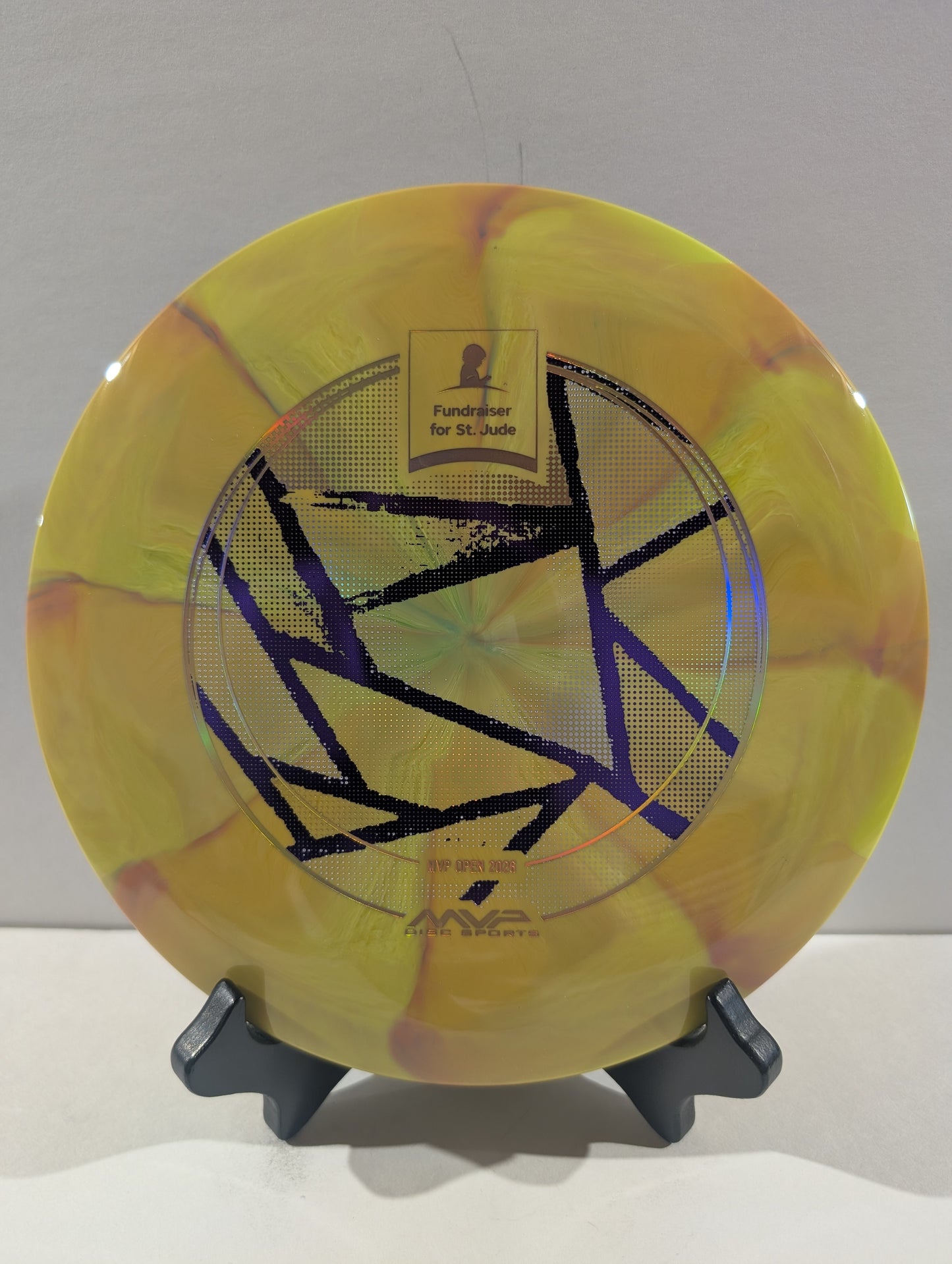A yellow disc golf disc with a purple geometric star design and a gold 'Foundation for St. Jude' patch, displayed on a small black stand against a light gray background.