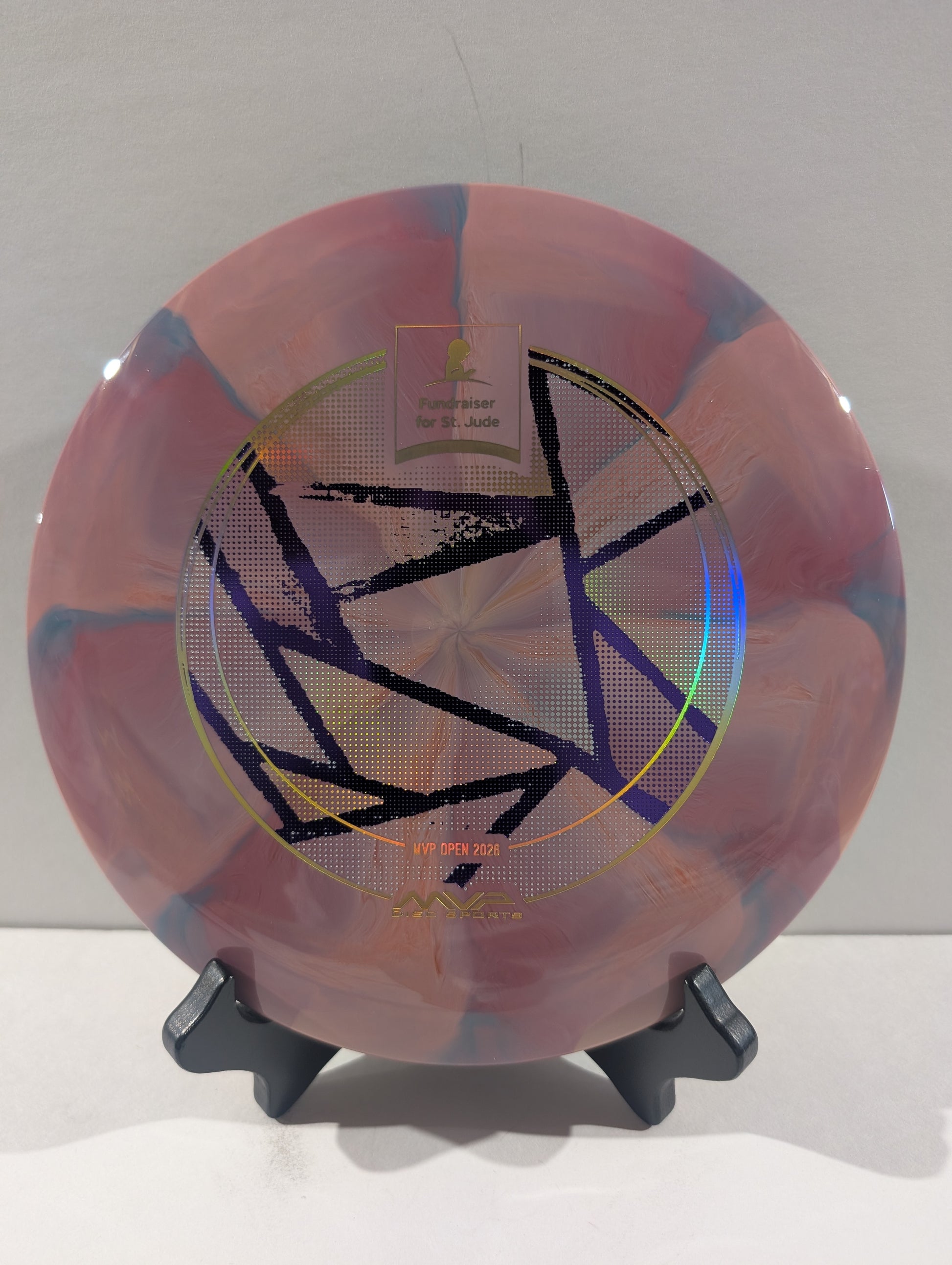 A disc golf disc with a pink, purple, and blue marbled pattern, featuring a black geometric star design and foil stamp in the center, displayed on a black stand against a neutral gray background.