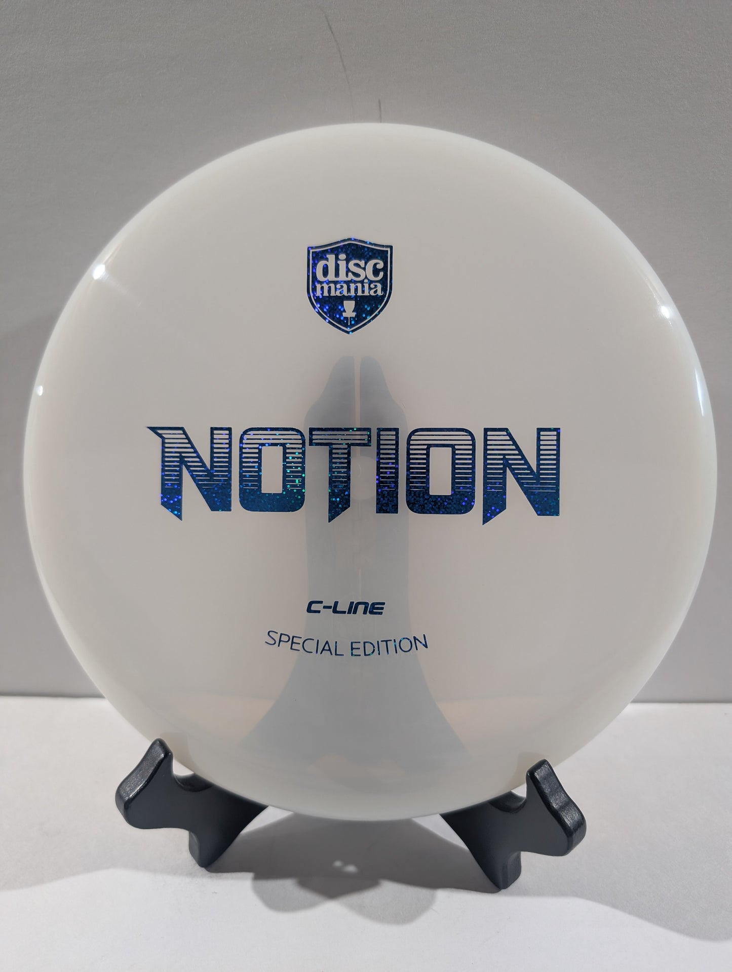 A white Discmania Notion C-Line Special Edition disc golf disc displayed on a small black stand against a neutral gray background.