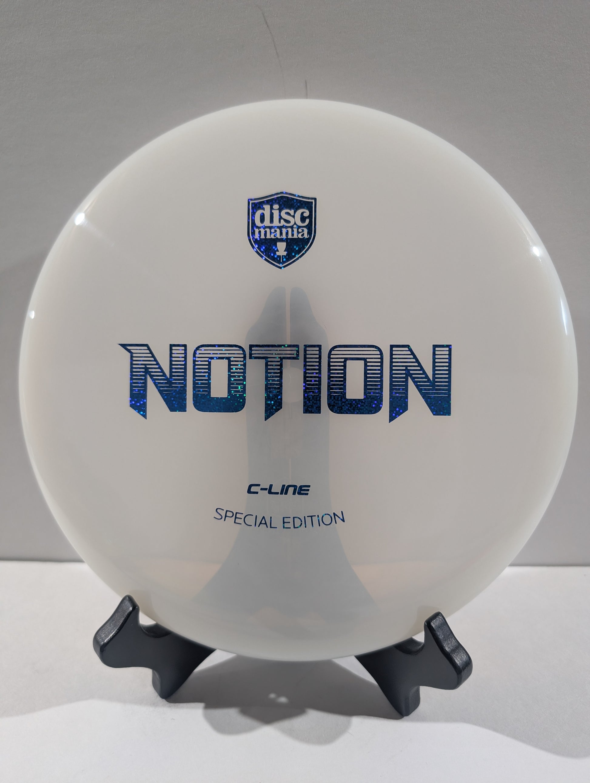 A white Discmania Notion C-Line Special Edition disc golf disc displayed on a small black stand against a neutral gray background.