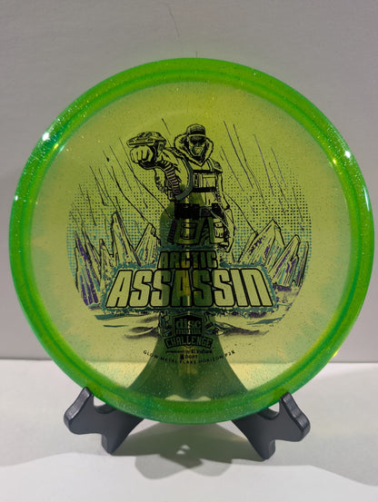 A Discmania Arctic Assassin disc golf disc with a green rim and a yellow center featuring a graphic of an Arctic soldier and mountains, displayed on a small stand.