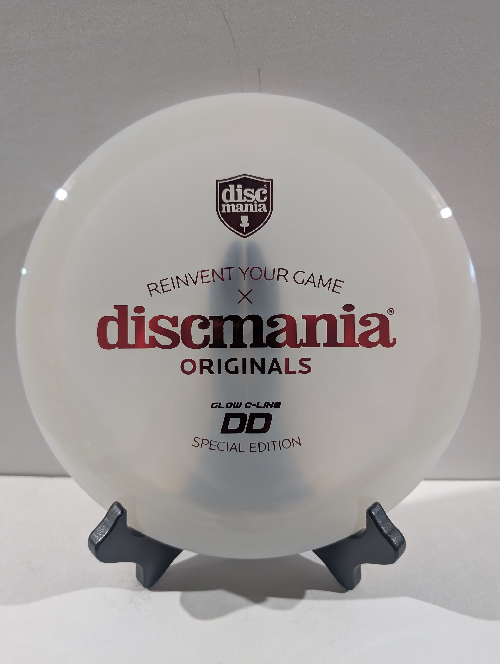 A white Discmania Originals DD Special Edition disc golf disc displayed on a small black stand against a light gray background.