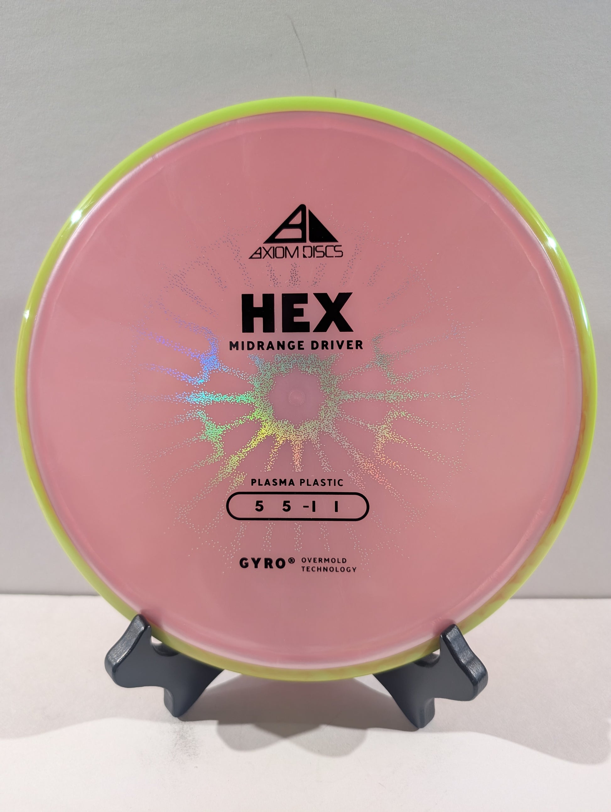 A pink disc golf disc with a yellow rim, displayed on a small black stand against a light gray background. The disc features black printed text including the brand 'Axiom Discs' and model 'HEX'.