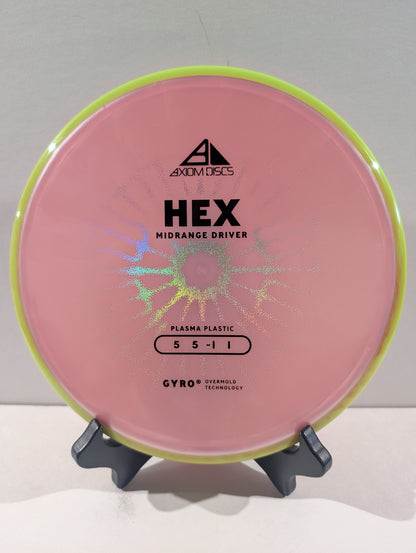 A pink disc golf disc with a yellow rim, displayed on a small black stand against a light gray background. The disc features black printed text including the brand 'Axiom Discs' and model 'HEX'.